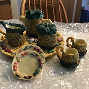 May include: A pineapple-themed tea set with a teapot, creamer, two teacups, and saucers. The set is decorated with green leaves, brown pineapple patterns, and colorful fruit accents.