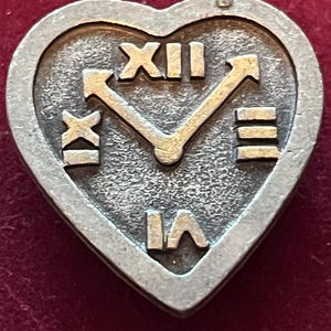Pode incluir: Um broche de metal prateado em forma de cora&ccedil;&atilde;o com uma superf&iacute;cie texturizada e algarismos romanos em relevo XII, III, VI e IX dispostos como um mostrador de rel&oacute;gio. Dois ponteiros de metal em relevo apontam para as posi&ccedil;&otilde;es XII e III.