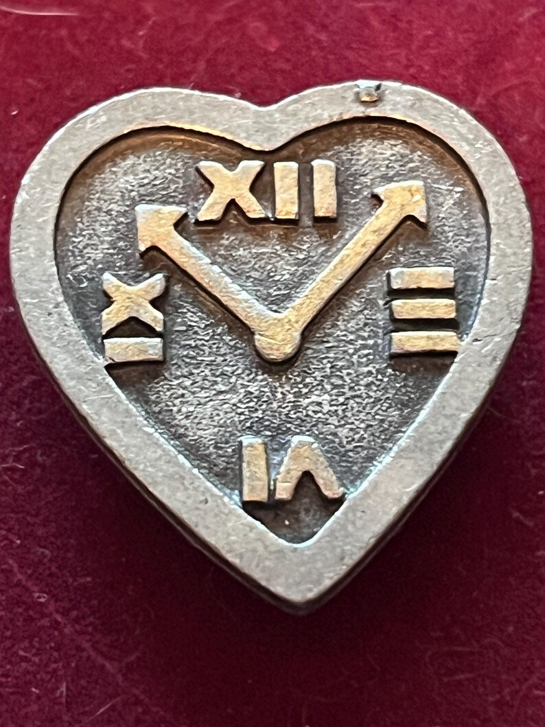 Pode incluir: Um broche de metal prateado em forma de cora&ccedil;&atilde;o com uma superf&iacute;cie texturizada e algarismos romanos em relevo XII, III, VI e IX dispostos como um mostrador de rel&oacute;gio. Dois ponteiros de metal em relevo apontam para as posi&ccedil;&otilde;es XII e III.