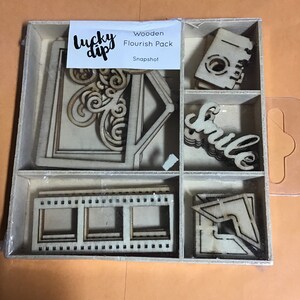 May include: Wooden craft kit with the words "Lucky Dip" and "Snapshot" on the packaging. The kit includes various wooden shapes, including a frame, a house, a swirl design, a film strip, a word "Smile", and a corner piece.