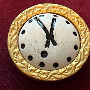 Pode incluir: Um bot&atilde;o redondo com um centro branco, ponteiros de rel&oacute;gio pretos e marcadores de hora em pontos pretos. O bot&atilde;o &eacute; emoldurado por uma borda de metal dourado texturizado com um padr&atilde;o de redemoinho em relevo na borda externa.
