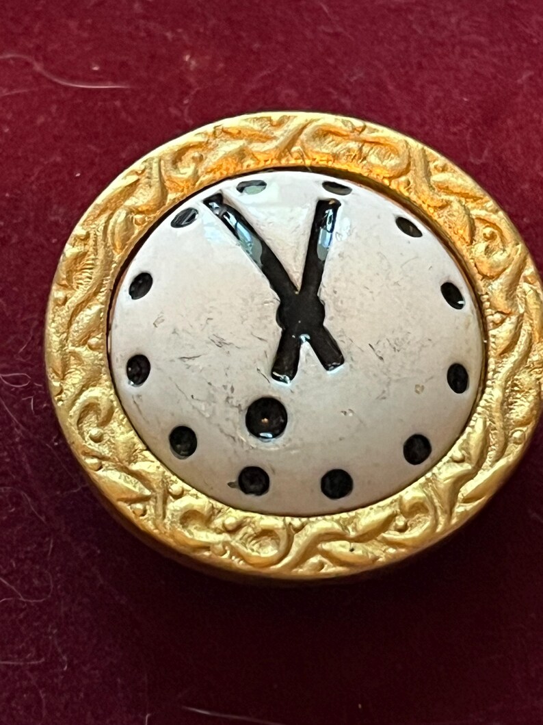 Pode incluir: Um bot&atilde;o redondo com um centro branco, ponteiros de rel&oacute;gio pretos e marcadores de hora em pontos pretos. O bot&atilde;o &eacute; emoldurado por uma borda de metal dourado texturizado com um padr&atilde;o de redemoinho em relevo na borda externa.