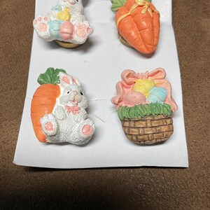 May include: Four Easter-themed ceramic pins. One pin features a white bunny holding a basket of colourful eggs. Another pin features a white bunny holding a carrot. The third pin features a carrot with green leaves and a yellow ribbon. The fourth pin features a brown wicker basket with green grass and colourful eggs.