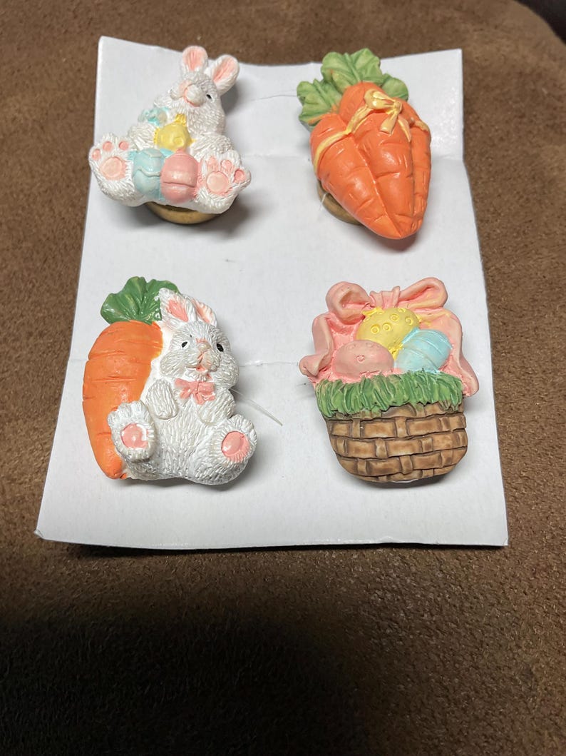 May include: Four Easter-themed ceramic pins. One pin features a white bunny holding a basket of colourful eggs. Another pin features a white bunny holding a carrot. The third pin features a carrot with green leaves and a yellow ribbon. The fourth pin features a brown wicker basket with green grass and colourful eggs.