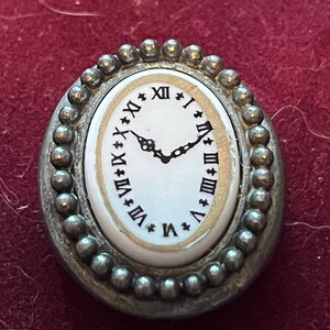 Pode incluir: Um broche de formato oval com um mostrador de rel&oacute;gio branco, algarismos romanos pretos e ponteiros pretos ornamentados. O centro &eacute; rodeado por uma borda de metal prateado texturizado com contas. O design imita um rel&oacute;gio de bolso.