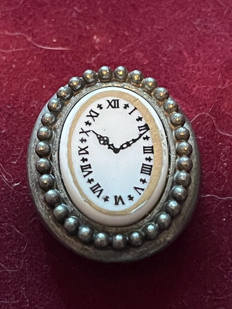 Pode incluir: Um broche de formato oval com um mostrador de rel&oacute;gio branco, algarismos romanos pretos e ponteiros pretos ornamentados. O centro &eacute; rodeado por uma borda de metal prateado texturizado com contas. O design imita um rel&oacute;gio de bolso.
