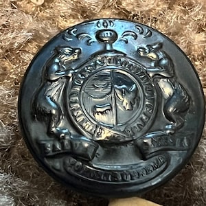 May include: A dark gray metal button with an embossed design. The button features a crest with two bear figures flanking a shield, and a banner with text. The button is circular and has a slightly raised edge.