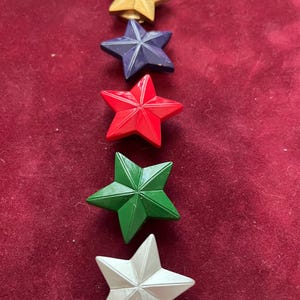 以下が含まれることがあります： 金、ネイビーブルー、赤、緑、白の星型の装飾品が5つ。星は赤い背景に垂直に配置されています。各星はテクスチャ加工された表面と3次元のデザインを持っています。
