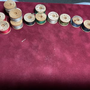 May include: A selection of vintage wooden thread spools in various colours, including red, blue, green, and silver. The spools are arranged on a burgundy surface. Some spools have printed labels with numbers and text.