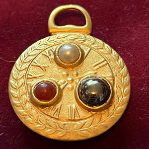 Pode incluir: Um pingente circular de tom dourado com uma borda texturizada em padr&atilde;o de folhas de louro. A face exibe algarismos romanos e tr&ecirc;s pedras caboch&atilde;o incrustadas: uma p&eacute;rola branca, uma pedra vermelho-alaranjada e uma pedra cinza-escura.