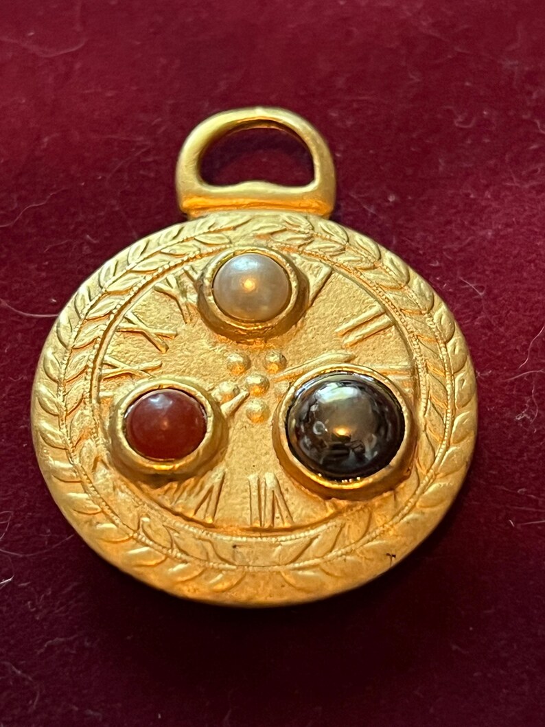Pode incluir: Um pingente circular de tom dourado com uma borda texturizada em padr&atilde;o de folhas de louro. A face exibe algarismos romanos e tr&ecirc;s pedras caboch&atilde;o incrustadas: uma p&eacute;rola branca, uma pedra vermelho-alaranjada e uma pedra cinza-escura.