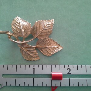 Könnte beinhalten: Goldfarbene Brosche aus Metall in Blattform mit drei Blättern und einem Stiel. Die Brosche ist etwa 5 cm lang.