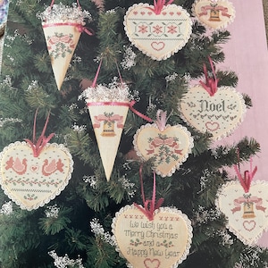 May include: A collection of cross-stitch Christmas ornaments featuring heart and cone shapes with lace trim and pink ribbon hangers. Designs include bells, holly, and the text "Noel" and "We wish you a Merry Christmas and a Happy New Year" on a printed page.