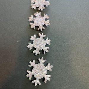 Könnte beinhalten: Vier weiße Glitzer-Schneeflocken-Ornamente in verschiedenen Größen, vertikal auf einem dunkelgrünen Hintergrund angeordnet.