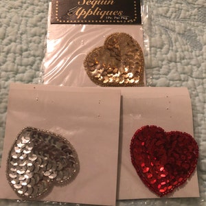 May include: Three sequin heart appliques in gold, silver, and red. The gold heart is in a package with the text "Sequin Appliques" and "Gold Heart". The other two hearts are on individual cards.