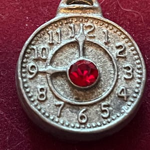 Pode incluir: Um pingente circular de metal prateado em forma de mostrador de rel&oacute;gio com numerais em relevo de um a doze. O centro apresenta uma &uacute;nica pedra preciosa vermelha redonda e facetada. Os ponteiros do rel&oacute;gio est&atilde;o fixos nas nove e doze.