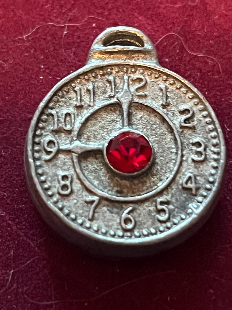 Pode incluir: Um pingente circular de metal prateado em forma de mostrador de rel&oacute;gio com numerais em relevo de um a doze. O centro apresenta uma &uacute;nica pedra preciosa vermelha redonda e facetada. Os ponteiros do rel&oacute;gio est&atilde;o fixos nas nove e doze.