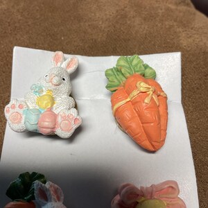May include: A set of four Easter-themed ceramic figurines. Two figurines are of white bunnies, one holding a yellow and blue egg and the other holding a carrot. The other two figurines are carrots with green leaves.
