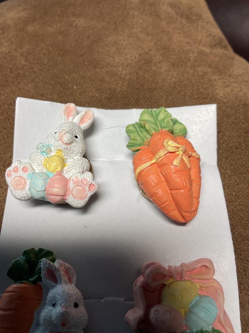May include: A set of four Easter-themed ceramic figurines. Two figurines are of white bunnies, one holding a yellow and blue egg and the other holding a carrot. The other two figurines are carrots with green leaves.