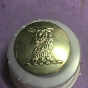 Könnte beinhalten: Ein goldfarbener Knopf mit einem erhabenen Bild eines Schafes. Der Knopf ist rund und hat eine glatte Oberfläche.