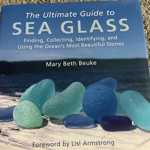 Może przedstawiać: Książka zatytułowana "The Ultimate Guide to Sea Glass" z kawałkami niebieskiego i zielonego szkła morskiego. Okładka zawiera tytuł, autorkę Mary Beth Beuke i podtytuł "Finding, Collecting, Identifying, and Using the Ocean's Most Beautiful Stones".