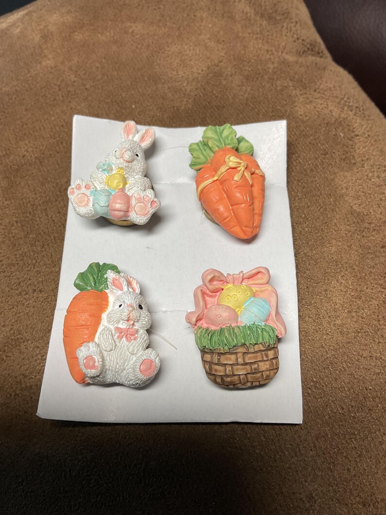 May include: Set of four Easter-themed ceramic buttons featuring a bunny holding an egg, a carrot, a bunny holding a carrot, and a basket of eggs.