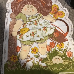 May include: A vintage fabric panel featuring a Cabbage Patch Kid illustration. The doll has brown hair, a floral dress, and is holding a watering can. The panel includes a butterfly, flowers, and the text "LEAVE OPEN FOR STUFFING".