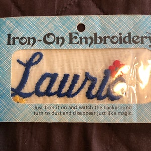 Puede incluir: Parche de bordado termoadhesivo con el nombre "Laurie" en escritura azul. El parche está sobre un fondo blanco y empaquetado en una tarjeta con estampado azul y blanco. La tarjeta dice "Iron-On Embroidery".