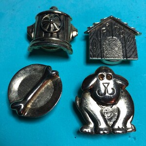 Puede incluir: Cuatro pines de metal de color dorado con diseños de temática canina. Los pines representan un hidrante, una caseta para perros, un hueso de perro en un plato y un perro de dibujos animados.