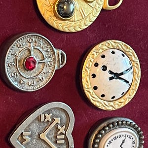 Pode incluir: Uma cole&ccedil;&atilde;o de cinco broches decorativos de metal em forma de rel&oacute;gios. O conjunto inclui um broche circular dourado com tr&ecirc;s pedras incrustadas, um broche circular prateado com um centro vermelho, um mostrador branco com borda dourada, um rel&oacute;gio prateado em forma de cora&ccedil;&atilde;o e um broche oval com borda de contas.