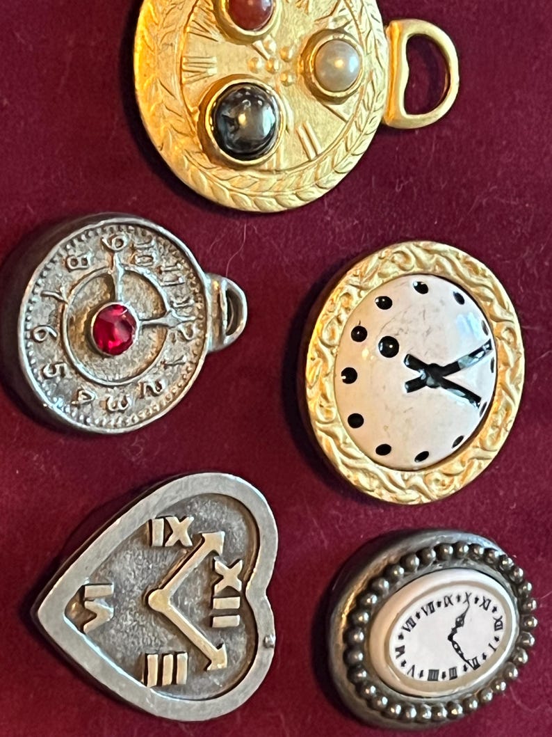 Pode incluir: Uma cole&ccedil;&atilde;o de cinco broches decorativos de metal em forma de rel&oacute;gios. O conjunto inclui um broche circular dourado com tr&ecirc;s pedras incrustadas, um broche circular prateado com um centro vermelho, um mostrador branco com borda dourada, um rel&oacute;gio prateado em forma de cora&ccedil;&atilde;o e um broche oval com borda de contas.