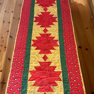 May include: A rectangular quilted table runner featuring a central gold panel with five red geometric star-like shapes. The runner is bordered by a dark green strip and outer red sections patterned with small gold stars and a gold edge trim.