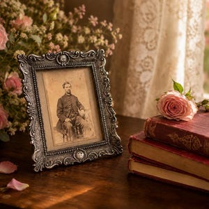May include: A rectangular picture frame with an ornate, dark metallic finish and intricate scrollwork detailing. The frame holds a sepia-toned vintage photograph of a person seated in a formal suit. The frame is positioned next to pink roses.