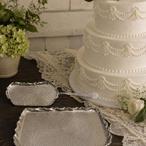 May include: A silver-toned metal cake server and a matching rectangular serving tray, both featuring a textured, radiating pattern on the surface and scalloped edges. The items are displayed near a three-tiered white cake with piped frosting.
