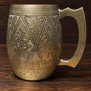 May include: A brass mug featuring an intricate engraved pattern of floral motifs and leaves arranged within diamond-shaped sections. The vessel has a smooth, flat rim and a sturdy, angular handle attached to the side, showcasing a metallic finish.