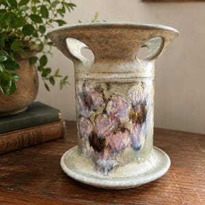 May include: A cylindrical ceramic oil burner with a wide flared top and base. The piece features a speckled cream and beige glaze with a prominent floral-like crystalline pattern in shades of soft pink, deep purple, and muted blue on the body.