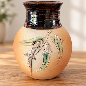 May include: A ceramic vase with a rounded body and a dark brown glazed neck. The light tan body features embossed relief designs of a lizard and stylised botanical elements with subtle green and white accents on the textured clay surface.