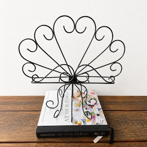 May include: A black metal cookbook stand featuring a decorative fan-shaped design with scrolled wire accents. The stand is positioned on top of a book titled AT OUR TABLE, which has a white cover with a colourful food photograph and black spine.