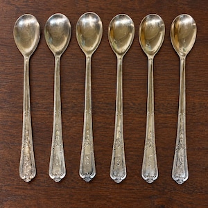 May include: A set of six silver-toned metal spoons arranged in a row. Each spoon features an elongated handle with a decorative embossed pattern along the edges and a rounded bowl. The metal has a polished finish with subtle signs of ageing.