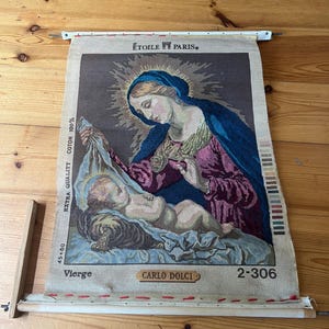 May include: A tapestry showing a religious scene with a woman in a blue veil holding a baby. The artwork features the text "ÉTOILE PARIS" and "CARLO DOLCI". The tapestry is mounted on a wooden frame and measures 45 x 60 cm.