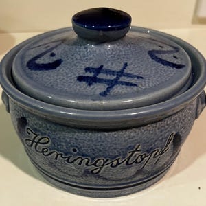 May include: A blue ceramic lidded pot with the word "Heringstopf" in cursive script. The pot has a textured surface and is decorated with dark blue abstract designs. The lid has a dark blue knob.