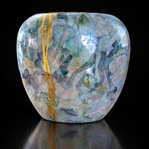May include: A rounded ceramic vase featuring an iridescent, watercolour-style finish in shades of blue, green, and white. A prominent, vertical, jagged gold-coloured line runs down the front surface, creating a striking contrast against the cool tones.