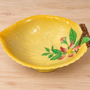 May include: A yellow ceramic bowl shaped like a leaf with a crackled glaze finish. The interior features a painted floral design with green leaves and pink petals. A brown textured handle resembling a tree branch is attached to the side.