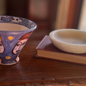 May include: A blue and white ceramic bowl with painted faces and a small, off-white stone bowl resting on a closed book. The ceramic bowl has a flared shape and is decorated with stylized faces.