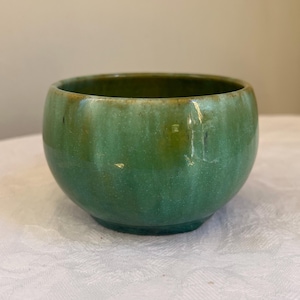 May include: A small, round, green ceramic bowl with a glossy finish. The bowl has a slightly flared rim and a dark green interior. The exterior colour transitions from a lighter green at the top to a darker green at the base.