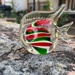 May include: A clear glass fish ornament with red, green, and white internal detailing. The fish has fins and a tail, and is resting on a textured grey surface. The ornament is a decorative item.