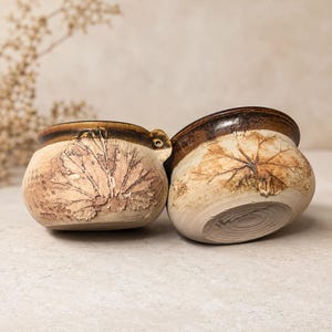 May include: Two round ceramic bowls with a light beige base and a dark brown glazed rim. Each bowl features a detailed, embossed leaf imprint on the exterior surface, showcasing natural textures and earthy tones in the clay finish.