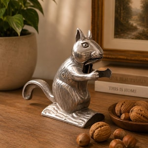May include: A silver-coloured metal nutcracker shaped like a squirrel sitting on a log base. The squirrel has textured fur details, pointed ears, and a mechanical jaw mechanism. A visible screw is located on the side of the squirrel's body.