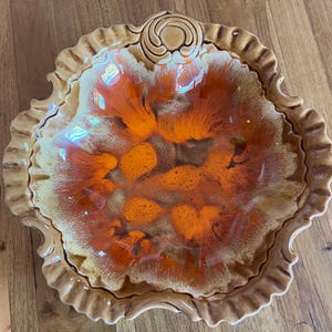 May include: A decorative ceramic serving dish with an unusual, organic shape. The dish has a brown exterior with a ruffled edge and a vibrant interior design of orange and brown hues. The dish has a diameter of approximately 30 cm.