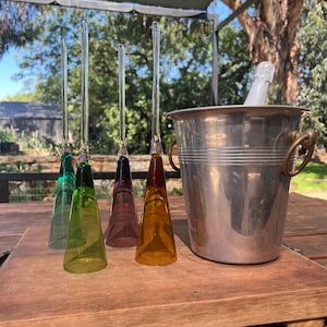 May include: Four coloured champagne flutes with long stems in green, yellow, purple, and orange are displayed on a wooden tray. A silver ice bucket with a champagne bottle is also present. The setting is outdoors.